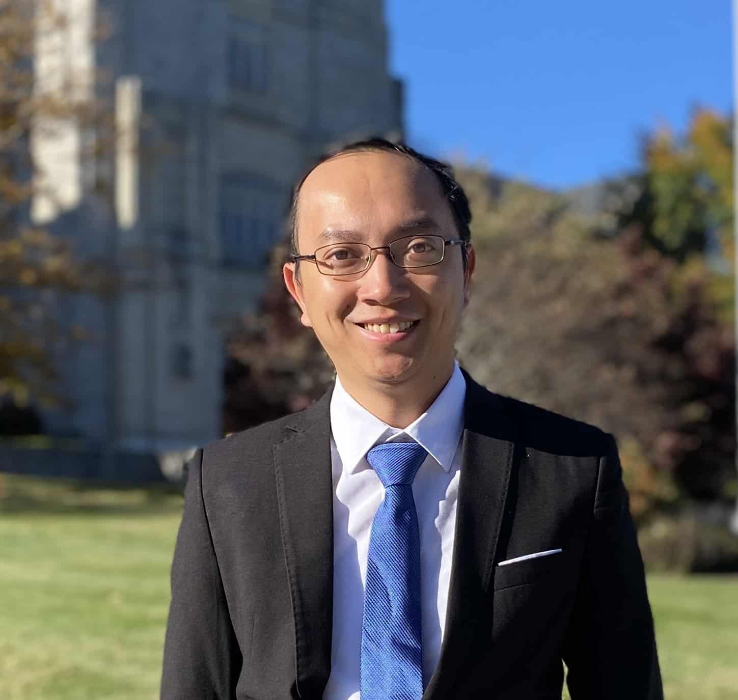 Man in dark suit, white shirt and blue tie standing in front of tall building and landscape