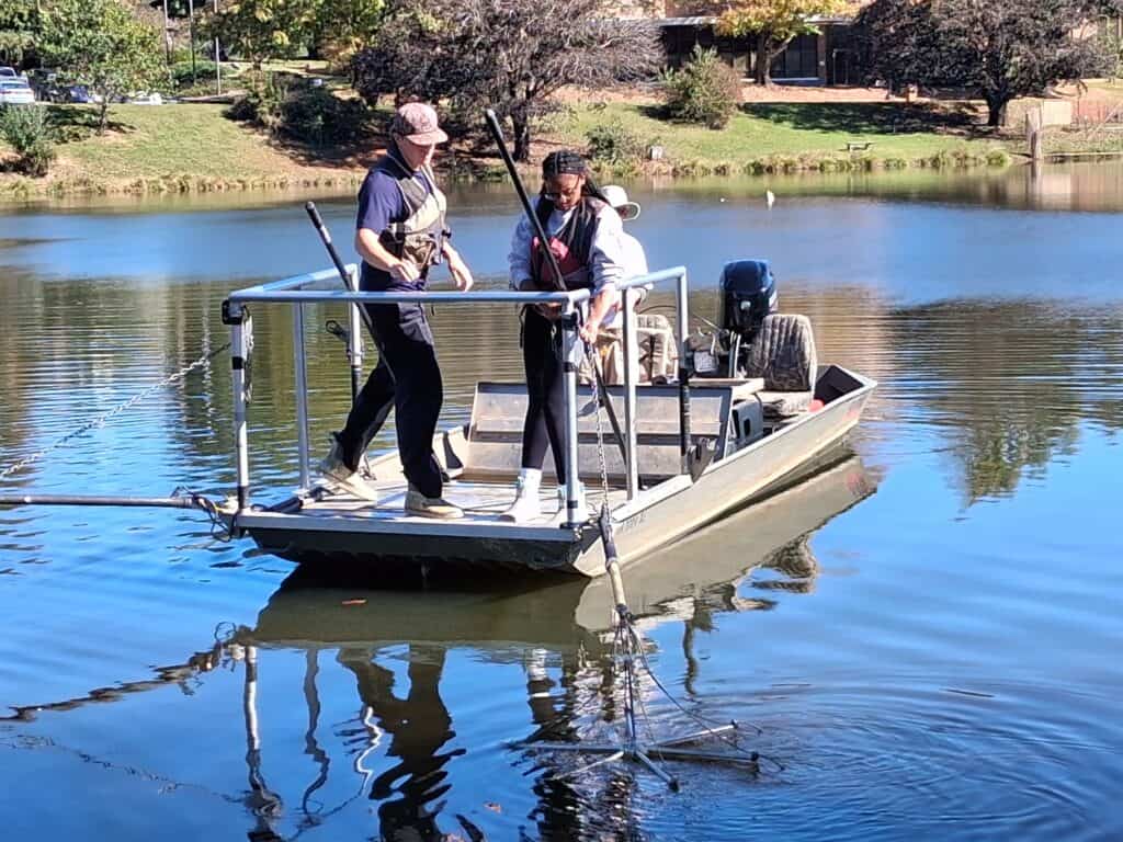 Two students wearing life vests are netting fish from a lake in a flat borttom boat being driven by a person in a hat