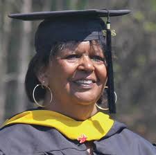 Head shot of Margaret Drakeford in black graduation cap and gown with gold collar, dark hair and big hoop earrings