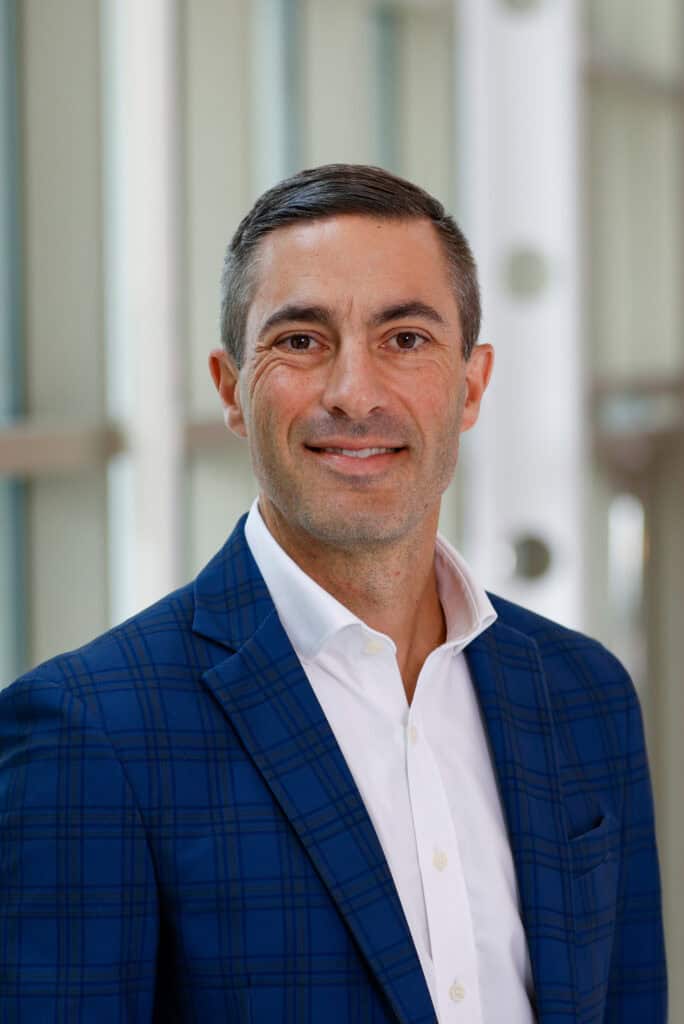 Head shot of Matthew Hubbard in dark jacket. white shirt, dark short hair, with muted white wall and doors or windows behind him