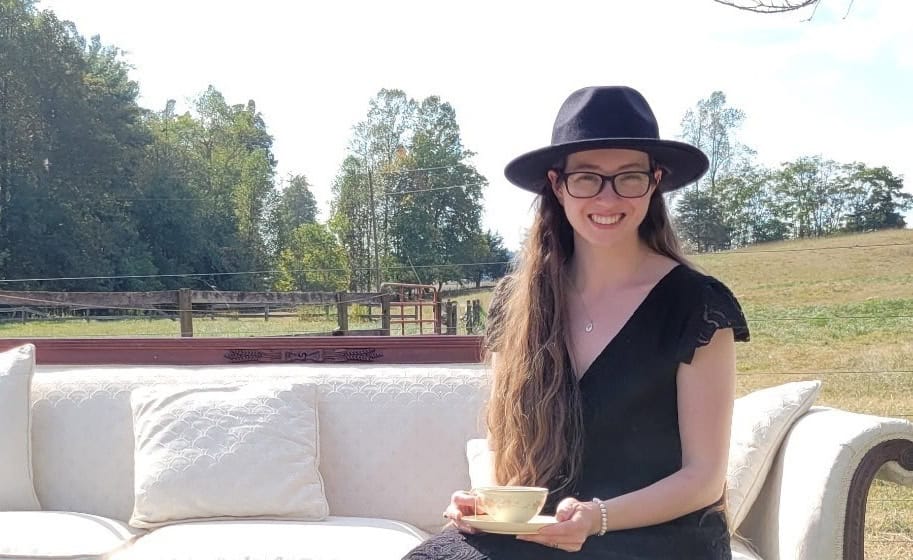 Senior Mattie Green sitting in a rural landscape on a white and dark wood vintage sofa in a long black dress with v-neck and tiny sleeves, with black hat and shoes, and a tea cup she's holding on her lap.