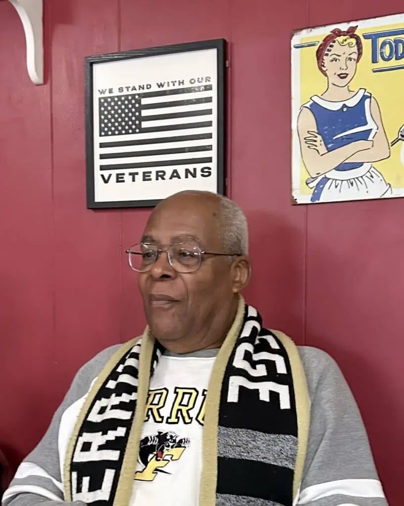 Dr. Melvin Mackin in Ferrum College clothing sitting under a sign about honoring veterans at the 77 restaurant in Ferrum