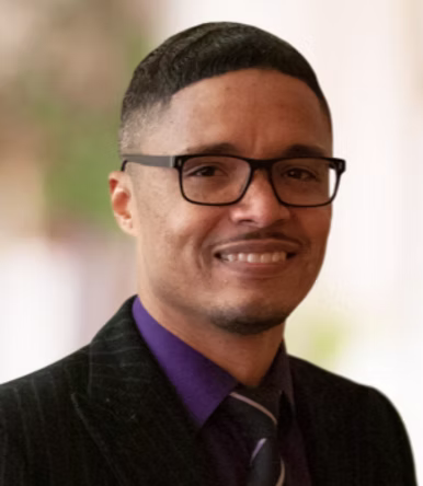 Headshot of man with short dark hair, dark glasses, dark shirt and jacket and tie, with a bit of muted color in background.
