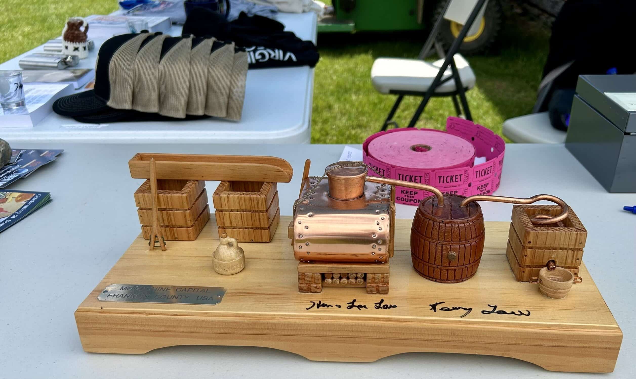 Miniature moonshine still in front of caps, shirts, and pink tickets at merch table at 2025 BRIM BBQ Cook Off event