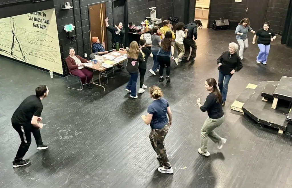 Actors in dark clothing circle around a black floor doing movement exercises, with 3 directors and managers between a table and a black wall in back, at The Rex Stephenson Theatre