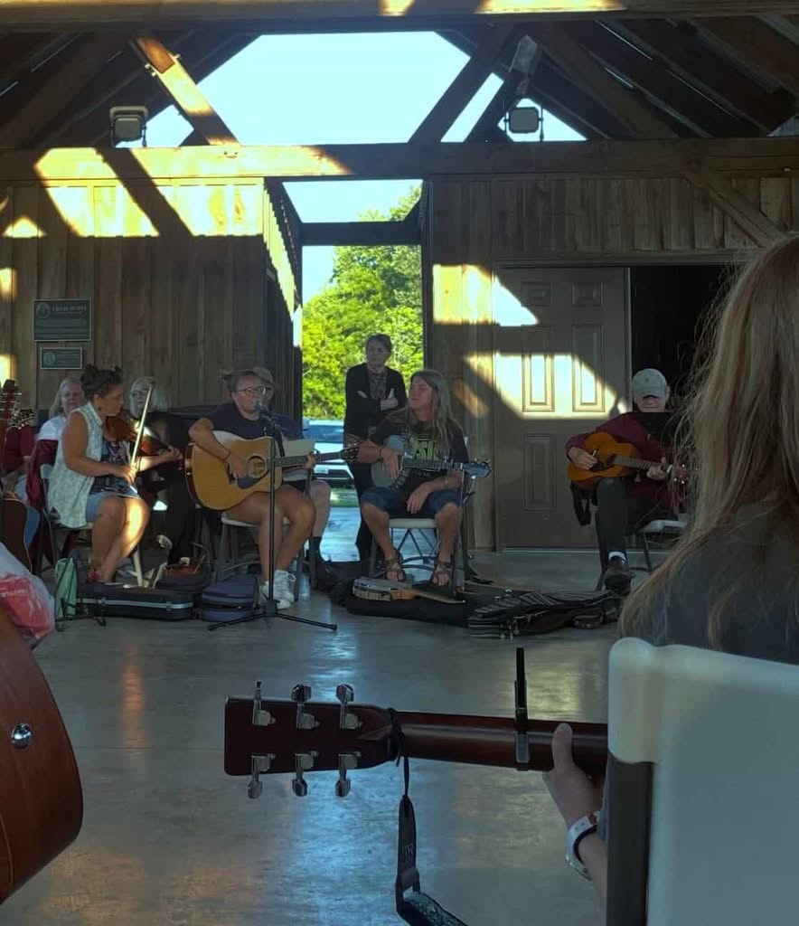Inside wooden farm pavilion in evening shadows. Seated musicians wearing summer clothes and one listener standing in shadows behind them.