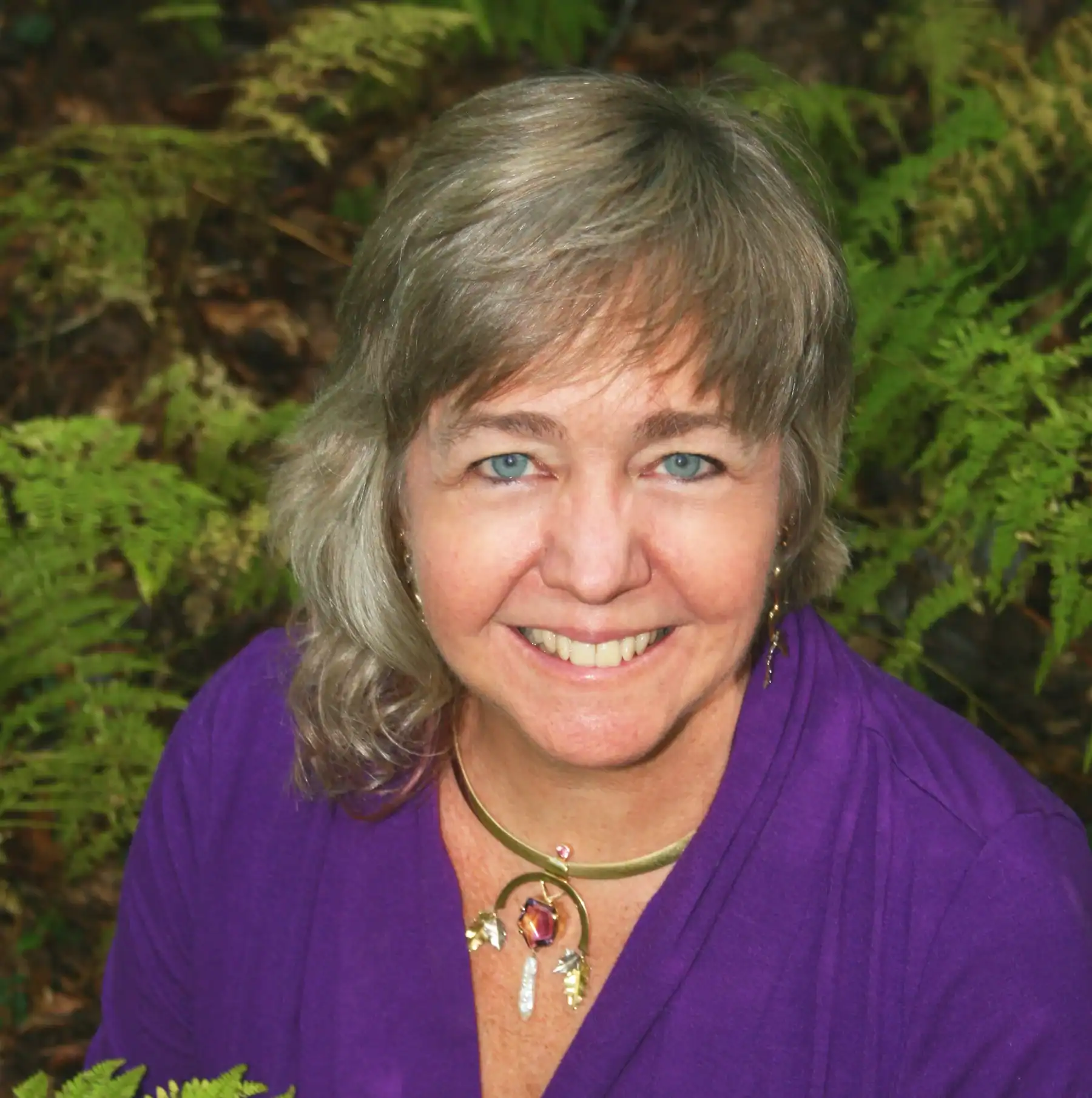 Headshot of Nell Fredericksen with purple shirt, multicolored necklace, green branches in background