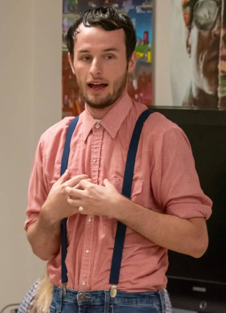 Student Jack Tale Player Nick Gore in red gingham shirt, blue suspenders, and short dark hair standing in front of posters with his hands in front of his chest