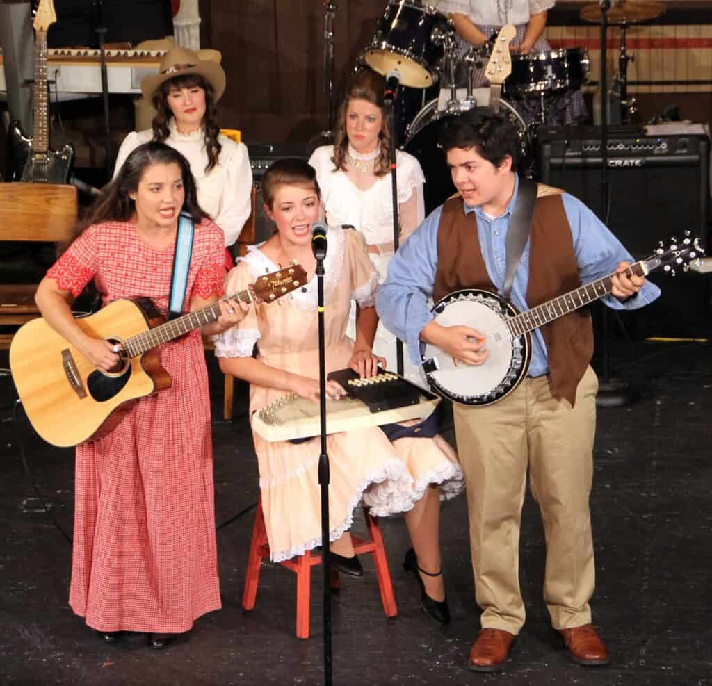 Five performers with country music instruments playing together on stage including Emily in long red print dress Rachel in back in white and one young man in brown vest and blue shirt