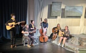 Rehearsal photo of Orchestra Appalachia, 6 people in black clothing and gold kerchiefs with their traditional old-time instruments on a theatre set with paintings of mountains above, blue background, and low structure painted like a mountaintop.