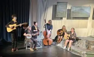 Rehearsal photo of Orchestra Appalachia, 6 people in black clothing and gold kerchiefs with their traditional old-time instruments on a theatre set with paintings of mountains above, blue background, and low structure painted like a mountaintop.