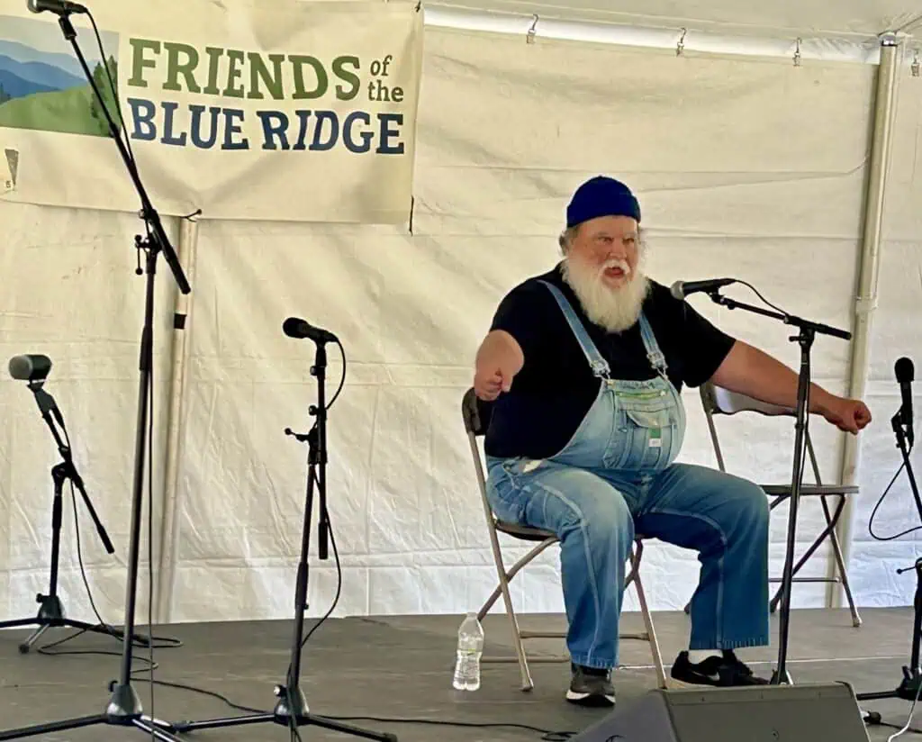 Orville Hicks in black shirt and overalls sitting in performance tent telling stories at Blue Ridge Folklife Festival