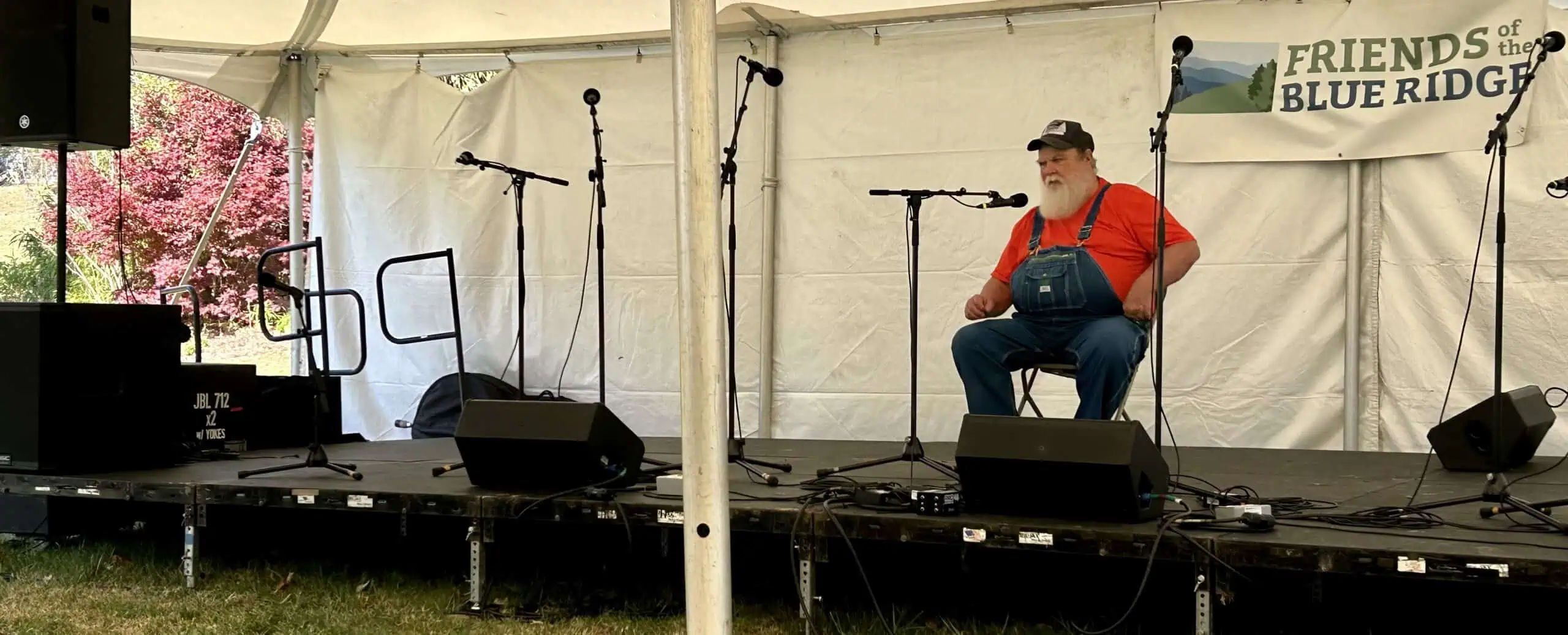 Orville Hicks seated in red shirt and overalls telling stories in performance tent at Blue Ridge Folklife Festival