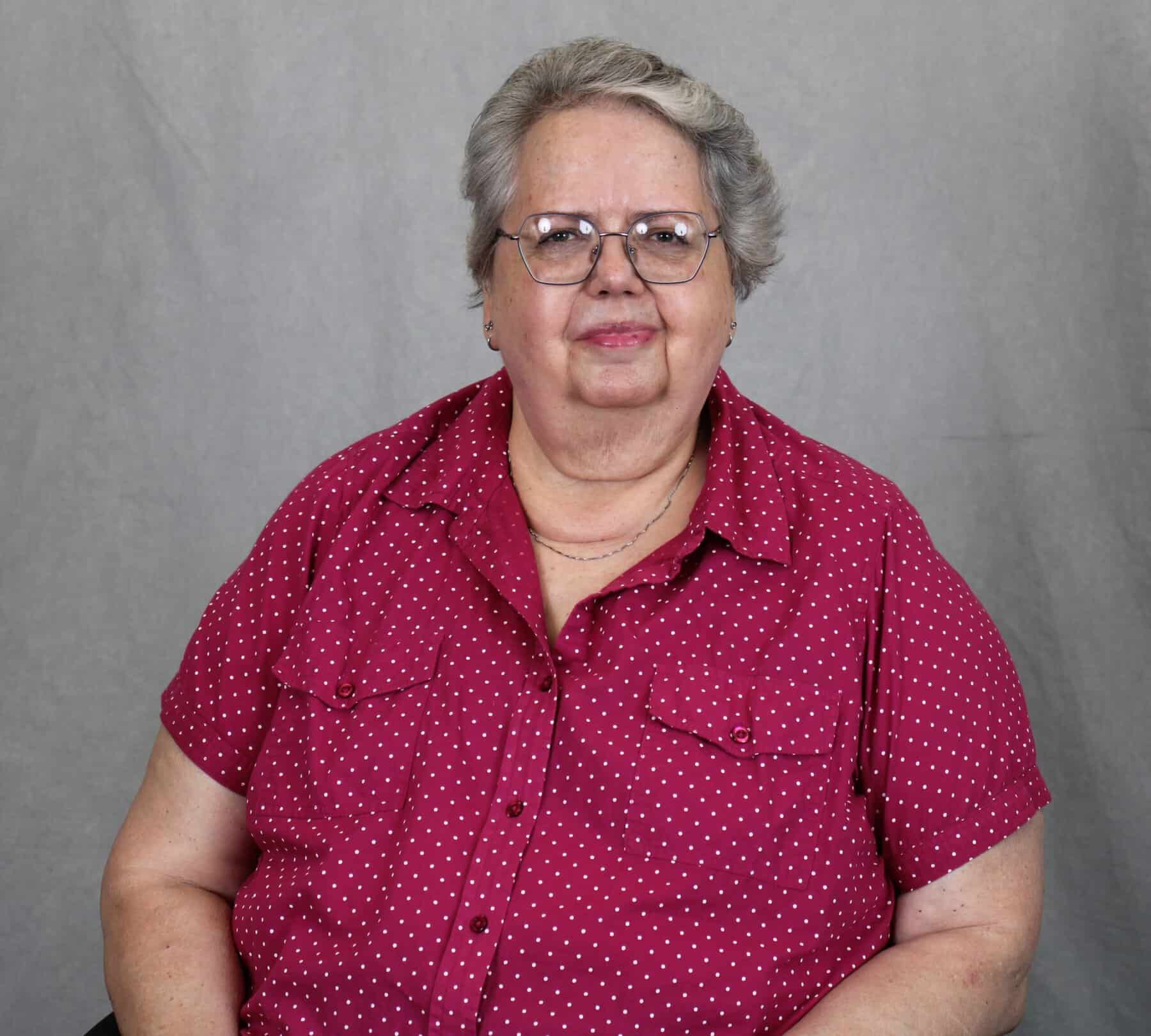 Headshot of Paula Geake in red short-sleeved shirt, necklace, glasses, and grey hair, with gray background