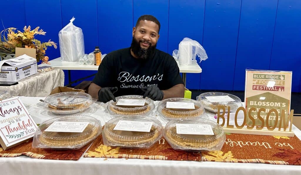 African American man with beard and dark shirt sitting behind a long table covered with pies he is selling. Blue wall and supplies are behind him.