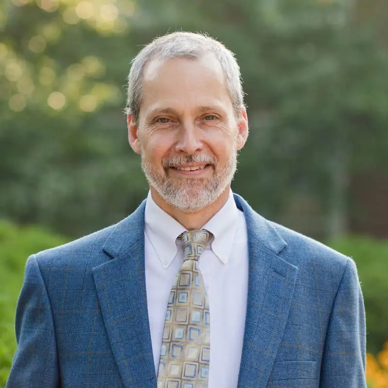 Head shot of Richard Grant with muted outdoor background, in blue jacket, white shirt and light patterned tie