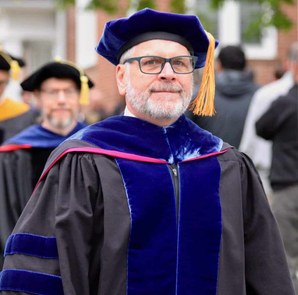 Karl Roeper in graduation line of faculty in black robe with velvet trim, red edging on blue velvet stoll, black tam with gold tassel, wearing glasses and grey facial hair
