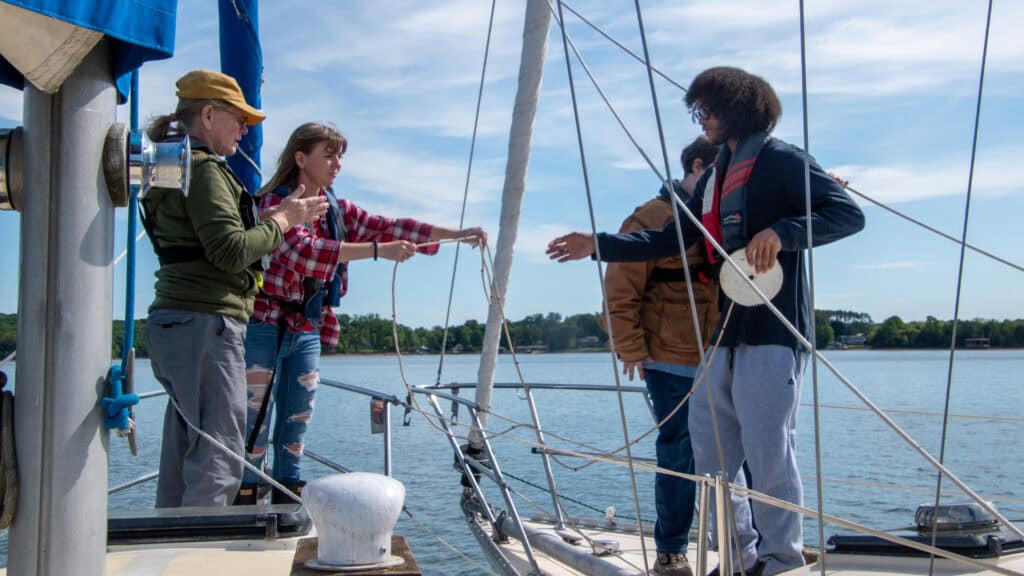 Student workers on a boat passing rope to each other. A woman is instructing them on what to do. The water is still and the sky has thin white clouds. There are trees and houses in the background. 