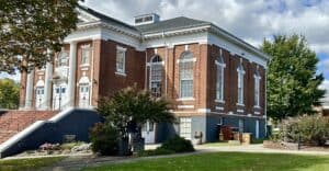 Schoolfield Hall, red brick with white trim, with tall front steps and large windows, bushes outside, and furniture on ground outside one of two side doors.