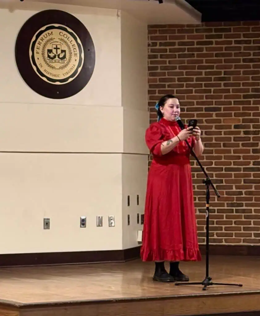 Scout Lynch in red dress reading from her writing in Panthers Den with Ferrum College seal on the wall