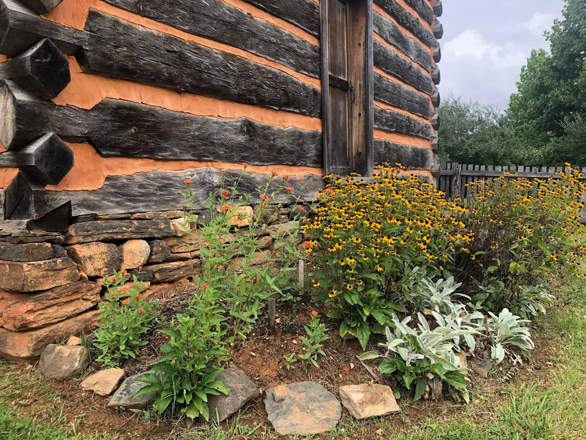 Portion of rustic wood and plaster farm museum wall with one wooden door and colorful flowers outside, with bits of trees in background