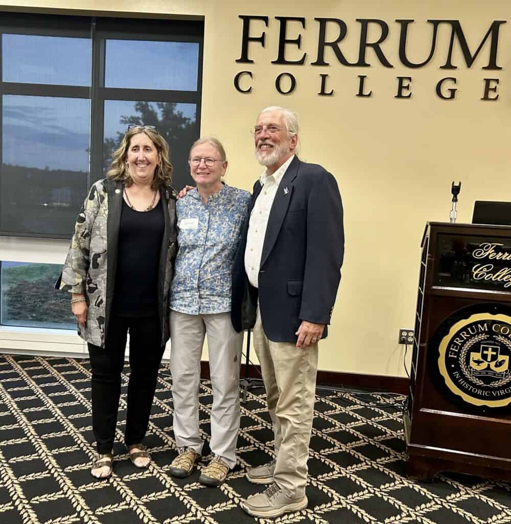Three adults standing close together for a recognition photo with Ferrum College words on gold wall and big window behind them: woman on left with dark hair and black clothes with black and gold and white patterned long jacket, woman in light blue flowered shirt and gray pants, and taller man with white hair and beard, glasses, dark jacket, and light shirt