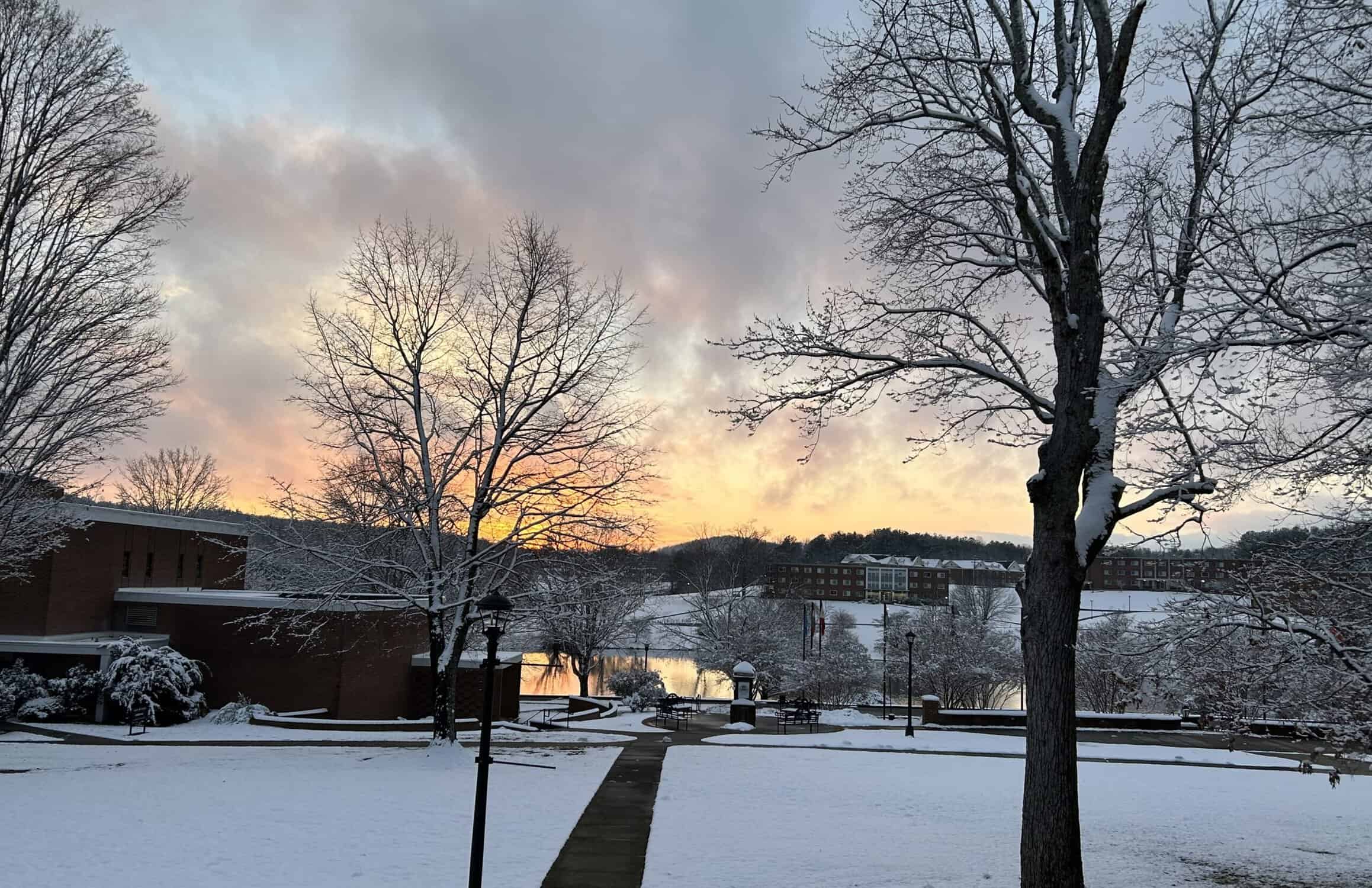 Central campus with snow on ground and trees, colorful sunset in mid-sky and on bits of lake showing in back, with sidewalk leading to brick buildings