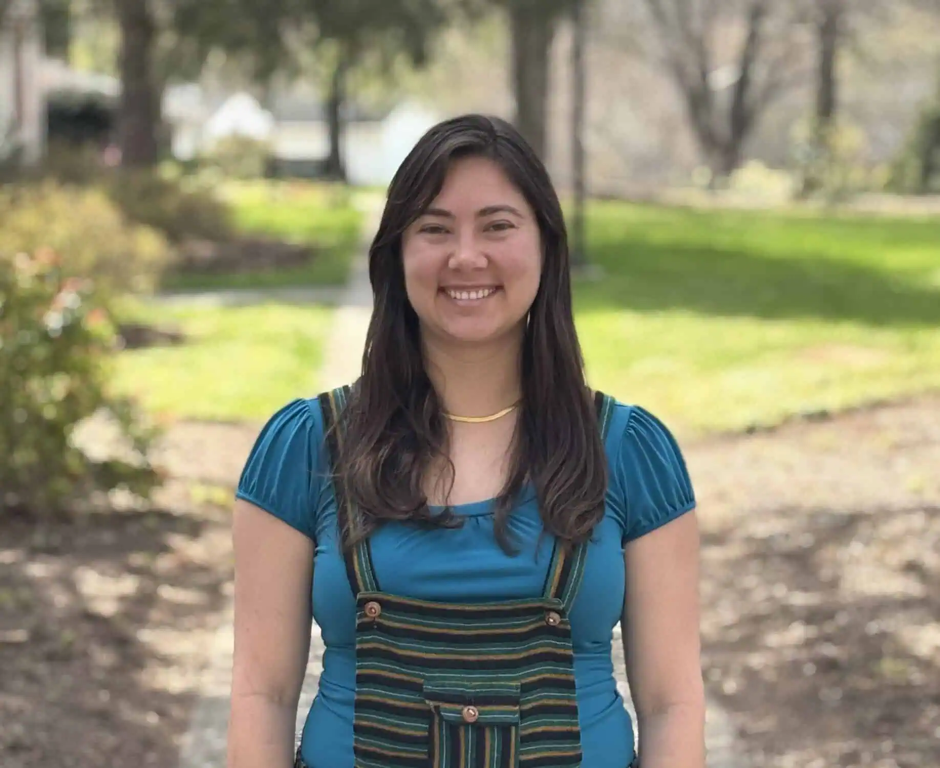 Sophia Fonseca head shot with long dark hair, teal short-sleeved shirt, and jumper with dark strips and straps, with a muted spring campus background