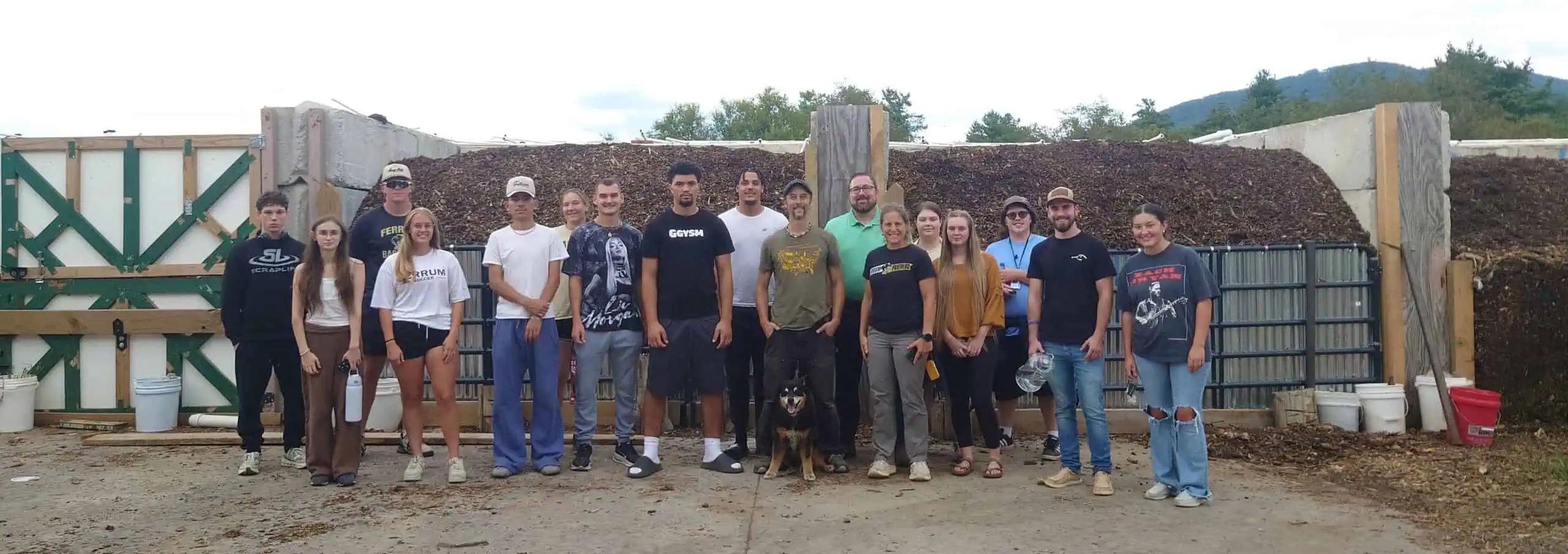 group of students staff and faculty stand in front of a row of compost bins