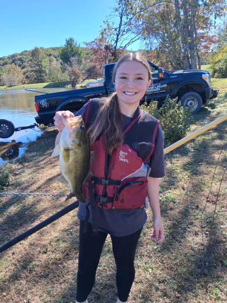 A female student wearing a life vest shows off a trophy bass with a truck, trees, and water behind her