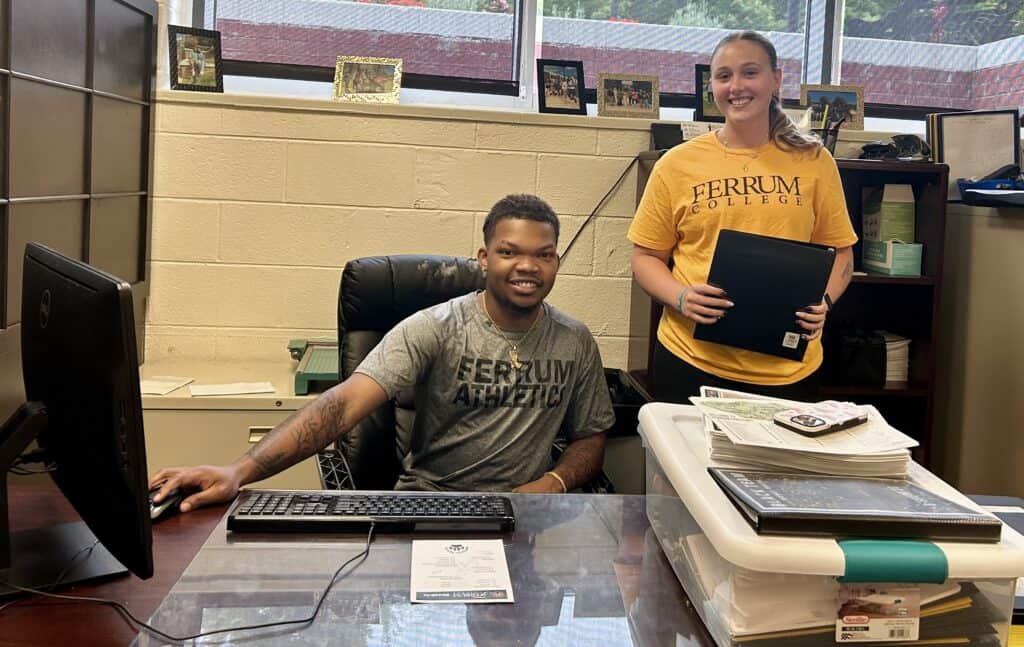 Two student workers behind a desk with a computer and office materials on it: a young man in grey shirt sitting and young woman in gold shirt standing with a black notebook. Behind them are a light brick wall and high windows.