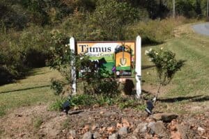 White Titmus Agricultural Center sign with colorful images, surrounded by plants, soil and trees in the background