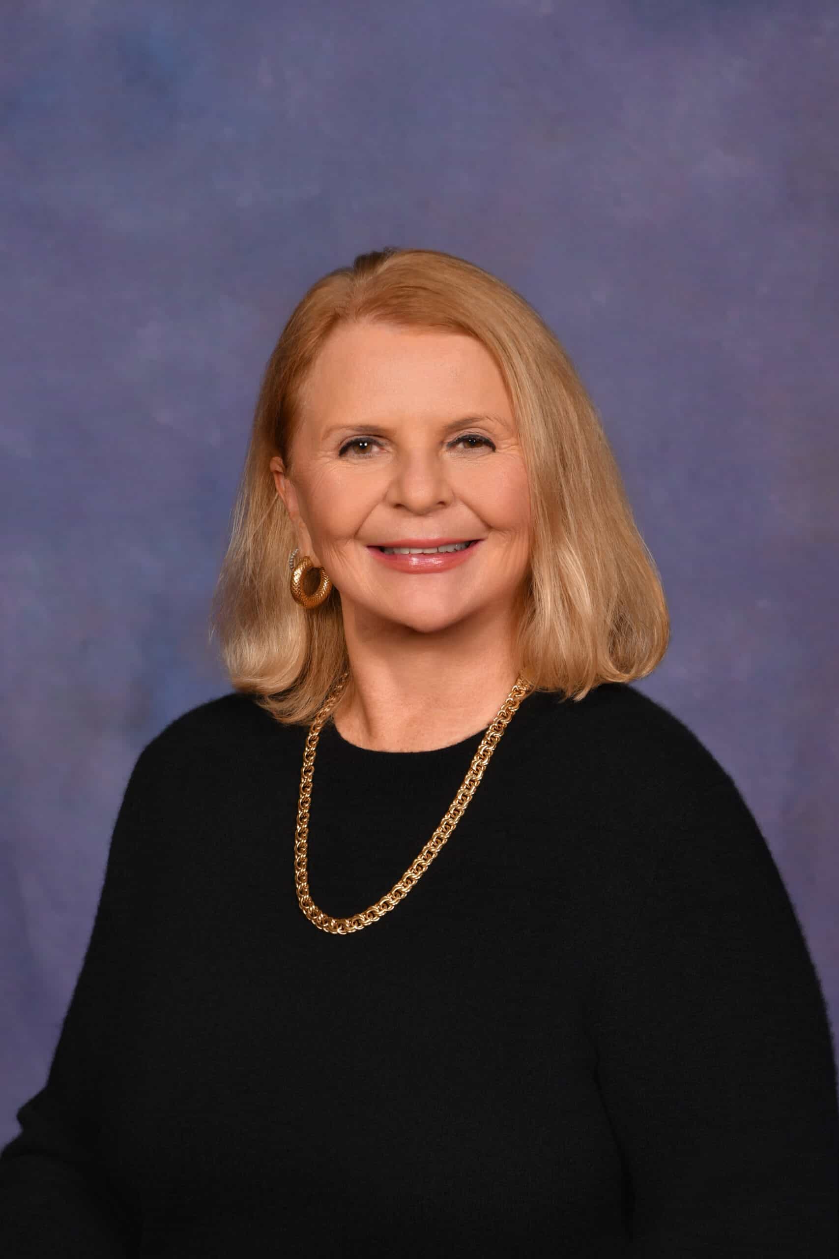 Toni Clark headshot--woman in black with a gold necklace, blond hair, against a dark background