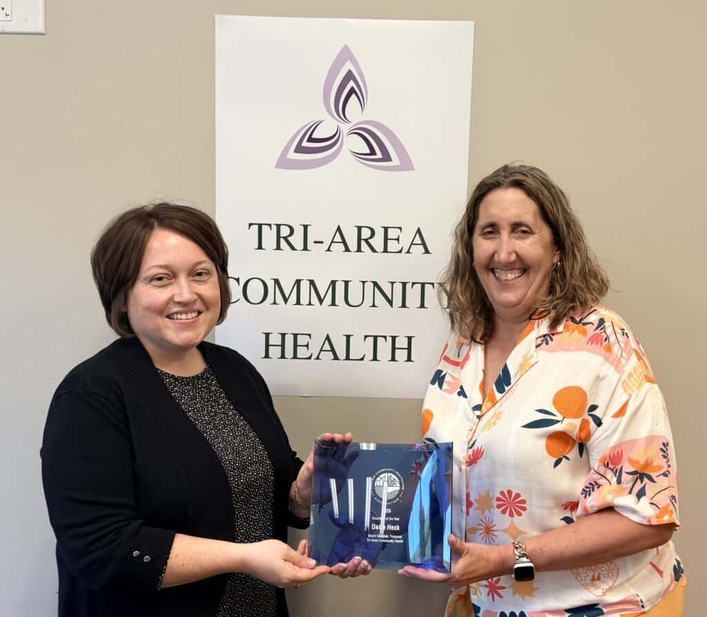 Delia Heck on right in dark hair and light flowered shirt receiving an award held between her and a woman with dark hair and clothes. Tri-Area Community Health name and triangular logo are on a sign behind them on a plain wall.