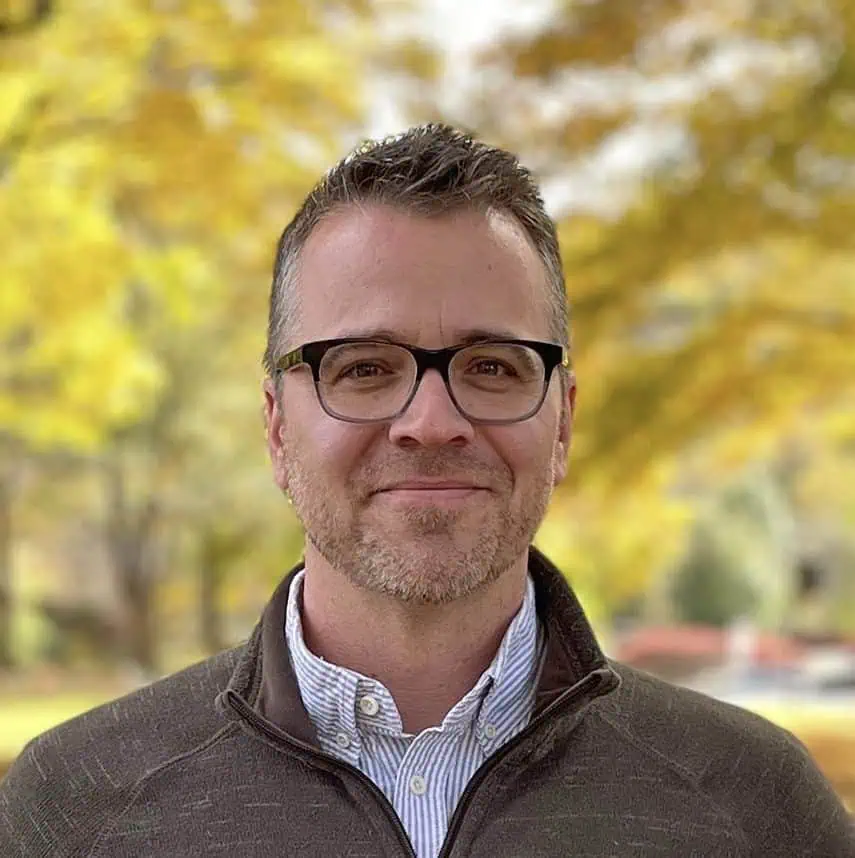 Headshot of Eric Vanden Eykel with blue shirt, brown jacket and glasses outside.