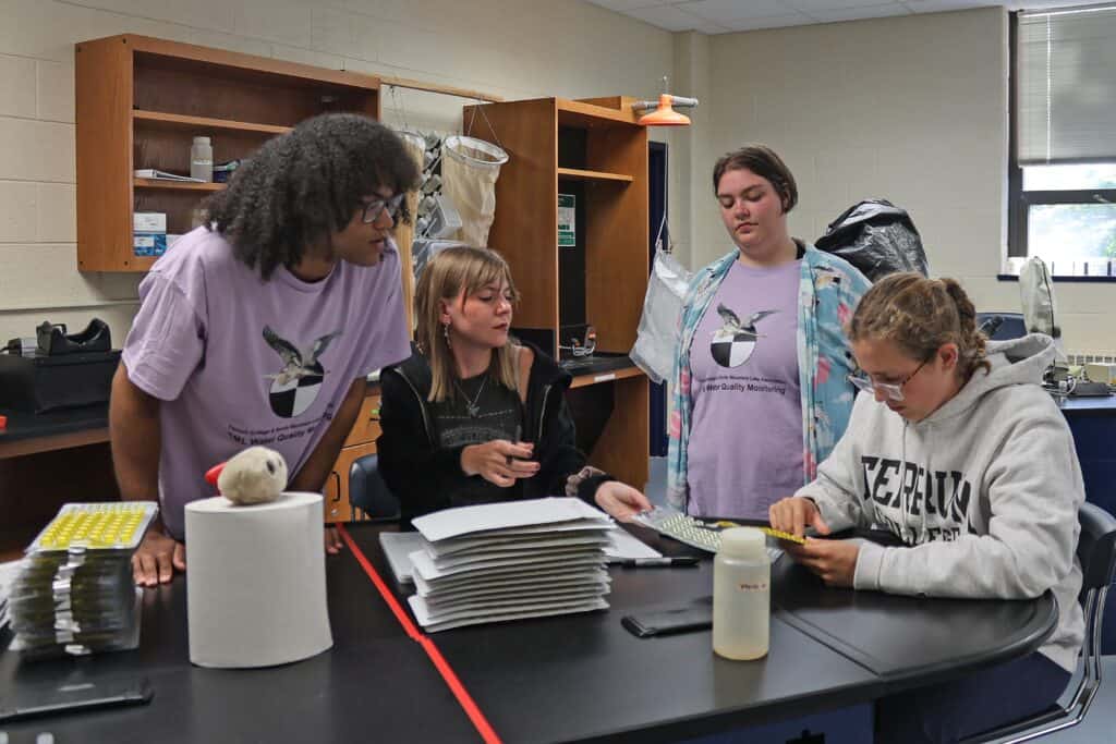 Four students working at a lab table with equipment and papers, wearing SMLA Water Quality t-shirts and Ferrum College sweatshirt