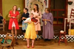 Scene from 2009 play "Wedding Belles" with four women in colorful dresses and one in yellow in front playing the violent in front of a front-porch setting