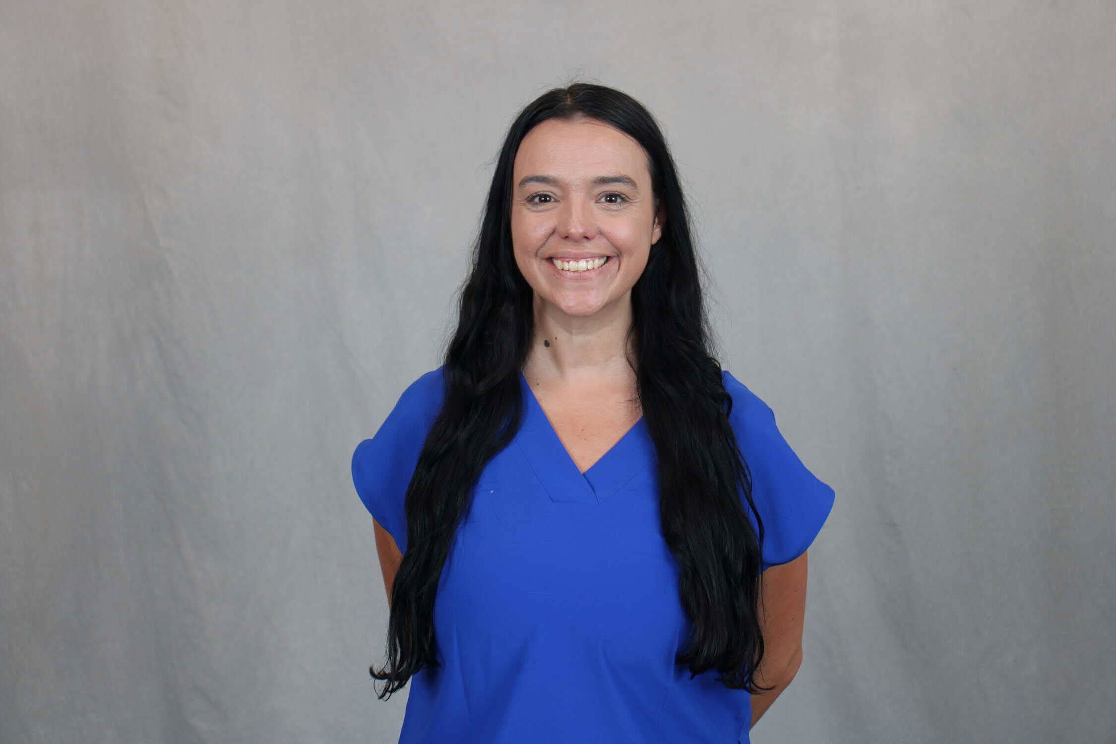 Head shot of woman in short-sleeved bright blue shirt, with long dark hair and grey background