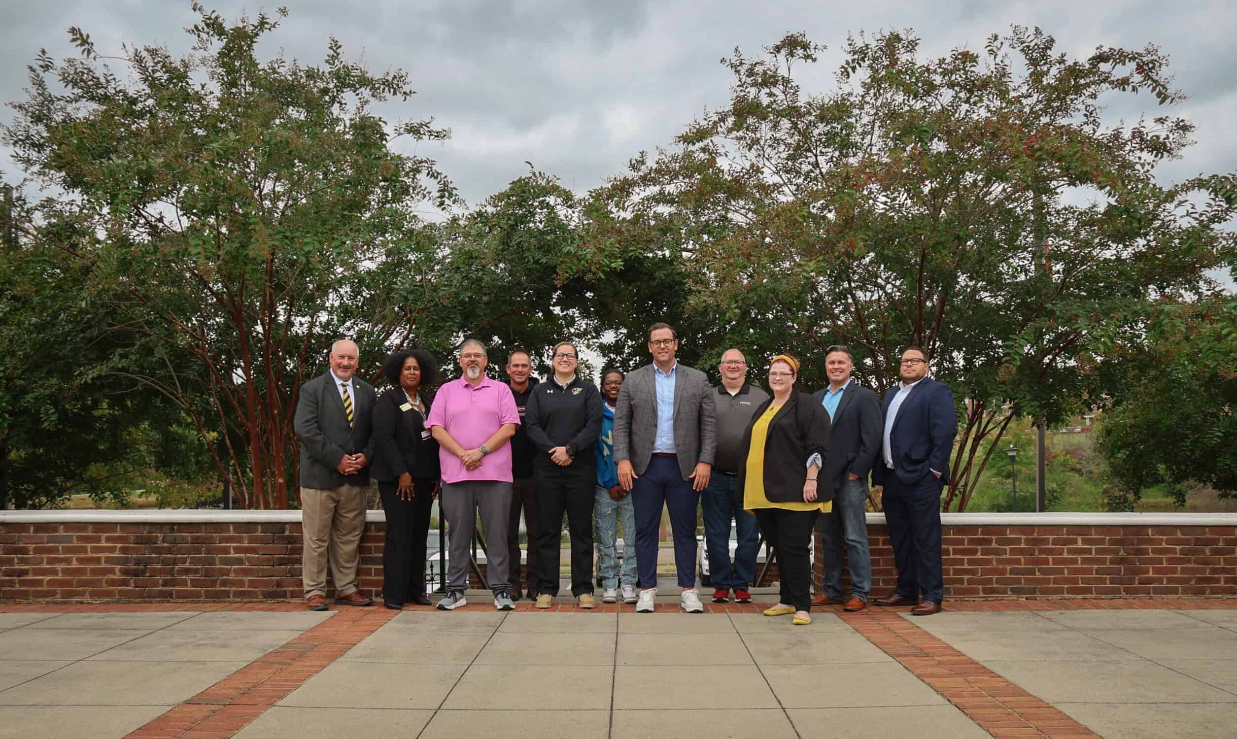 Alumni Board of Directors, a dozen adults lined up in front of a low brick wall on a brick plaza, with tall trees behind them.