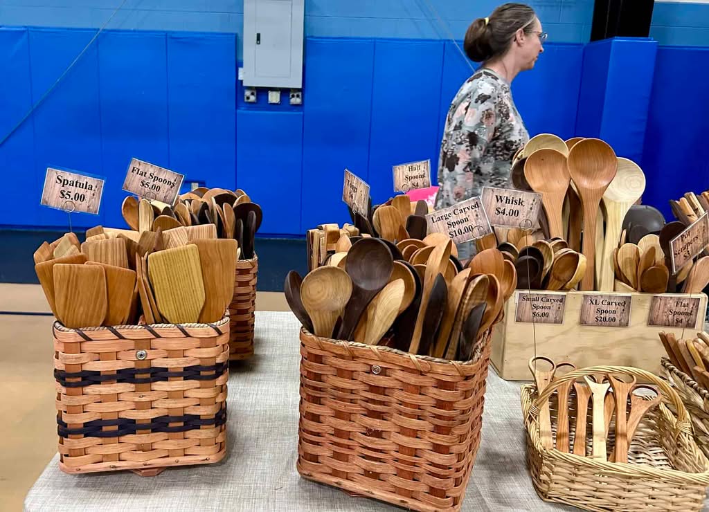 Different kinds of wooden spoons displayed on a table with white tablecloth. Blue wall and woman walking by in background