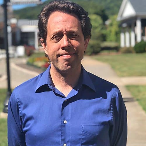 Headshot of Ferrum College professor Ed Hally standing outside on campus in a blue shirt with short dark hair