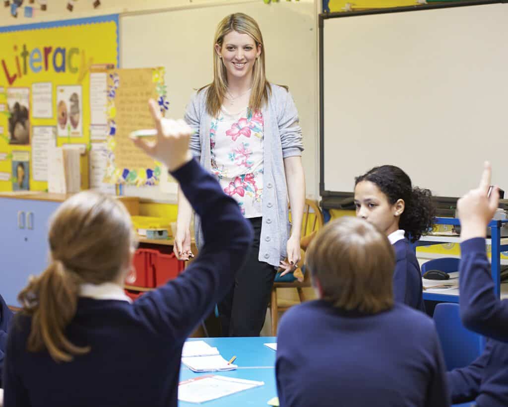 A teacher stands around a group of students, who are raising their hands to answer a question.