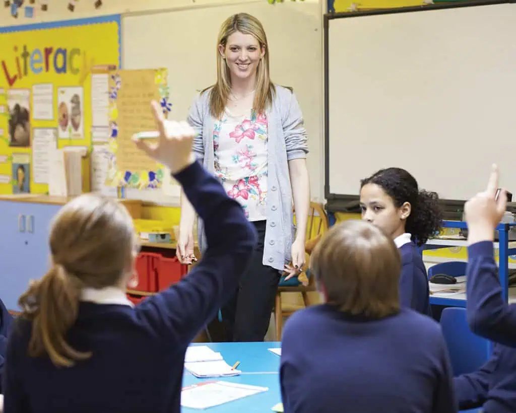 A teacher stands around a group of students, who are raising their hands to answer a question.