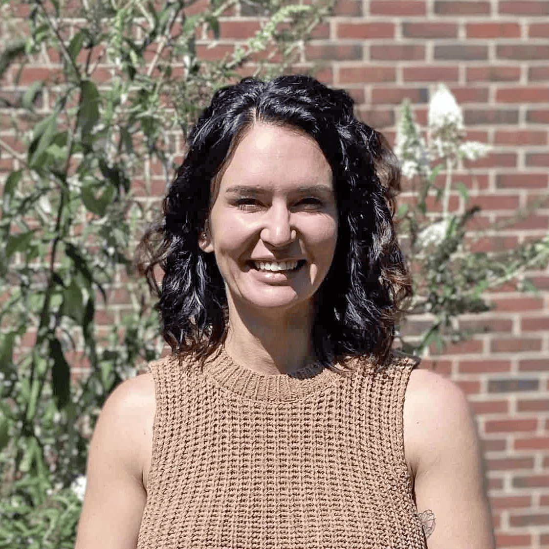 Photo of Abigail Jamison taken outside in front of a brick wall with a flowering bush behind her