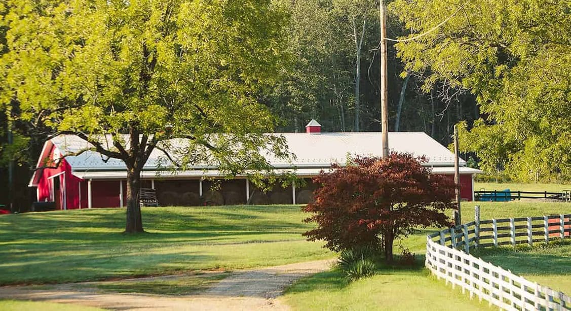 Red barn at Titmus Agricultural Center surrounded by treen trees and grass, brown bush, and some white fence showing