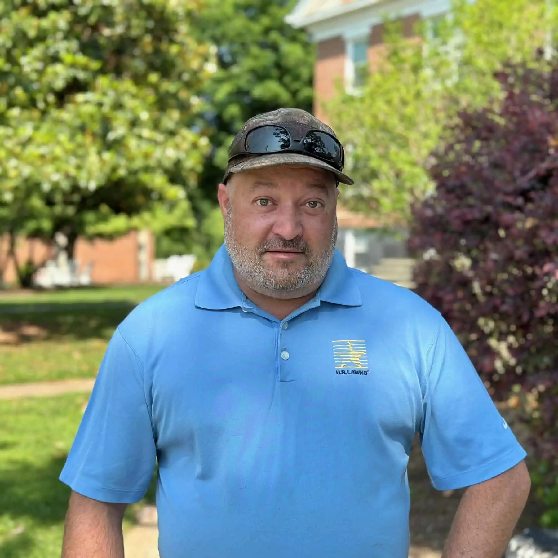 The photograph is of Andrew Bowles wearing a ball cap and sunglasses on his head and a blue polo shirt taken outside on a pretty summer day