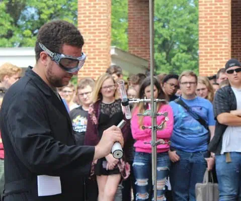 male professor in safety goggles presenting chemical reaction outside while other students are watching.