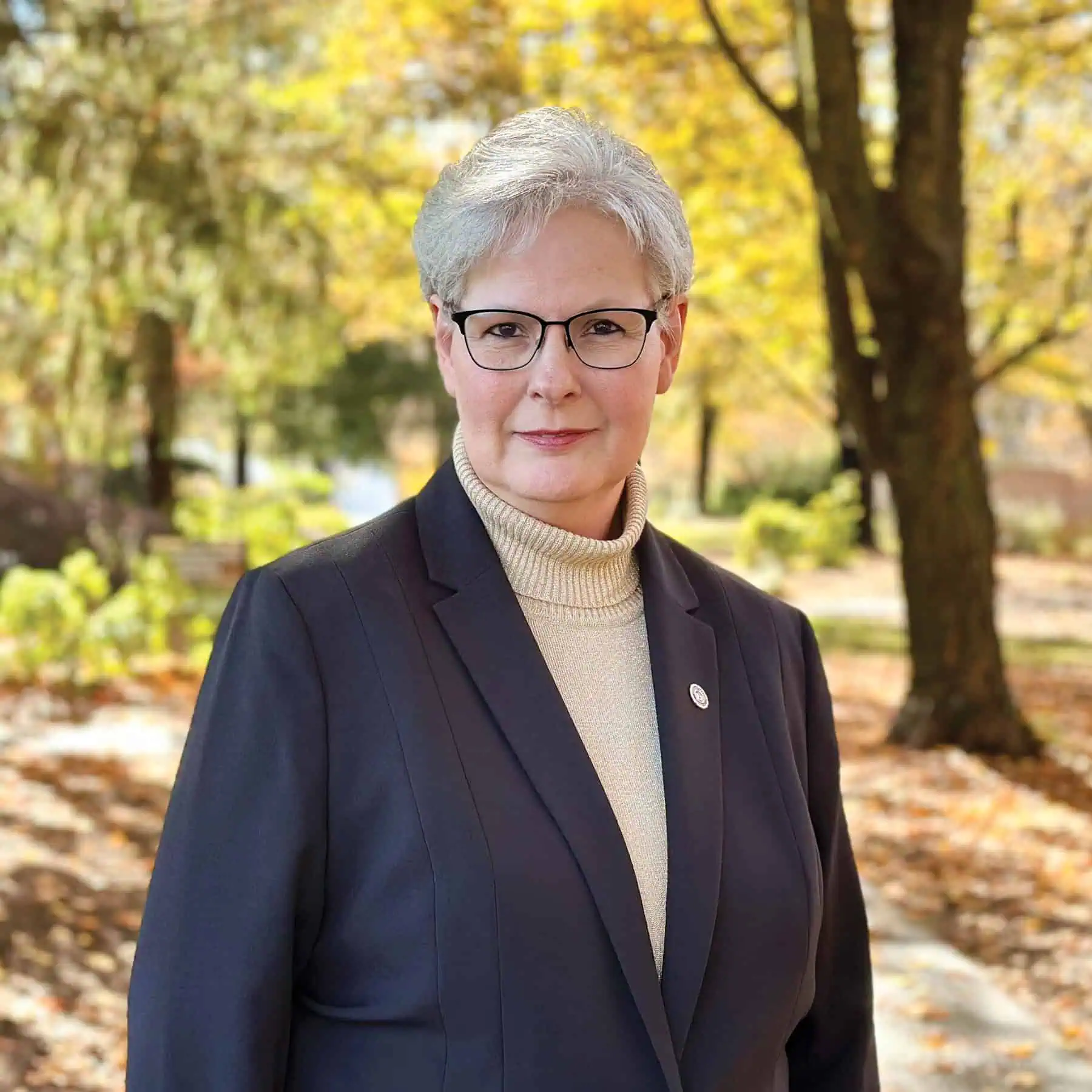 Photo of a woman with short gray hair, wearing glasses, a tan turtleneck, a black jacket, and gold lapel pin with trees and fall leaves in the background