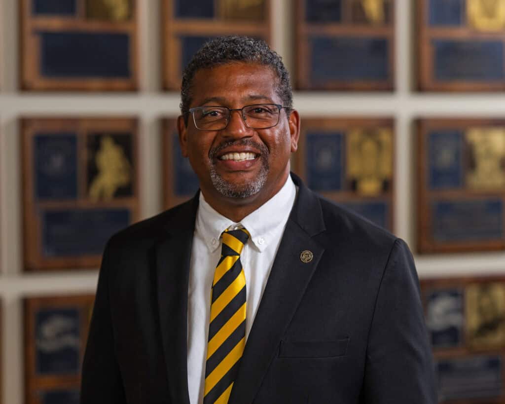 Cleive Adams with short dark hair, glasses, dark suit, white shirt, black and gold tie, in front of a muted wall of athletic plaques