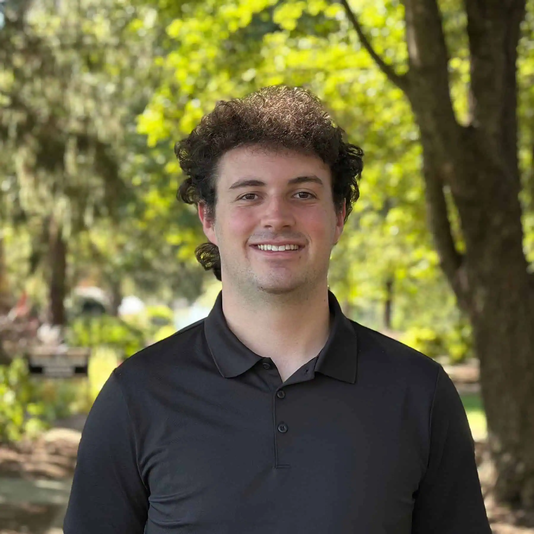 Young man with curly brown hair wearing a black polo shirt standing outside on campus on a pretty summer day